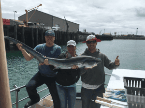 Three people proudly holding a large fish at a harbor.